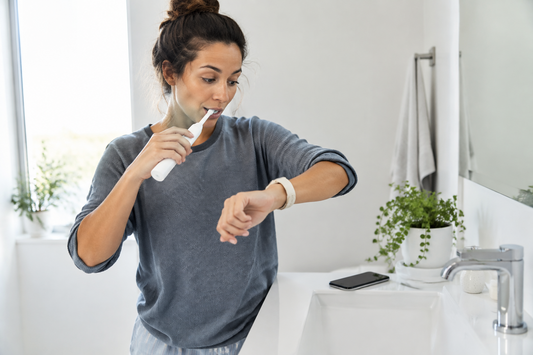 Person brushing teeth and checking time at bathroom sink — why brushing always comes first when you have no time to do both.