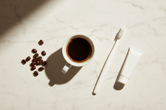 A white espresso cup with coffee beans beside a toothbrush and whitening tube on marble — coffee stains vs teeth whitening explained.