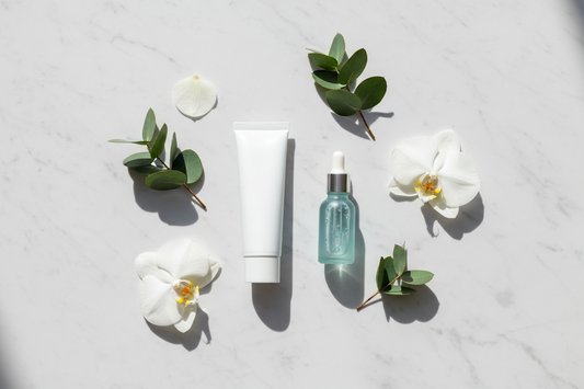 A minimalist flat lay featuring a white toothpaste tube, a skincare serum dropper bottle, botanical leaves, and white flower petals on white marble — representing the crossover of skincare ingredients like niacinamide into modern oral care.