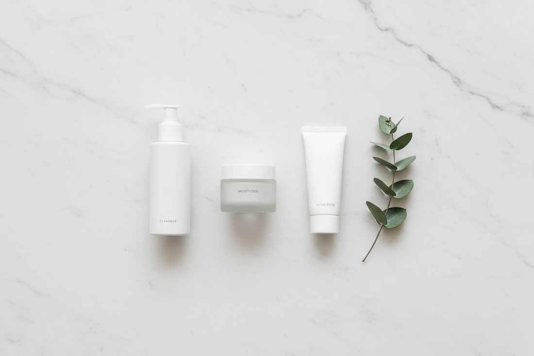 A clean top-down shot of three minimal skincare products — cleanser, moisturiser, and sunscreen — arranged in a row on white marble with generous white space and a single botanical stem, representing a simple, intentional 3-step skincare routine.